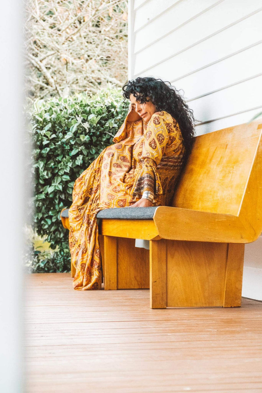 Woman in a yellow patterned dress sitting on a wooden bench outdoors.