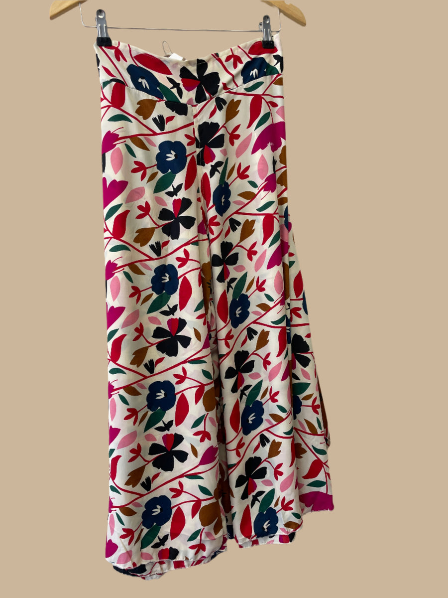 Wide-legged summer pants with an abstract floral design in navy, fuchsia, rose, red, black, and golden brown colors, displayed on a hanger with a decorative lamp and a mobile phone.